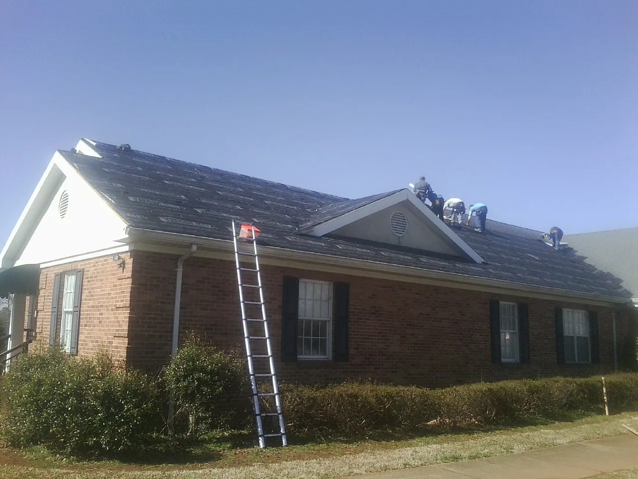 Crew tearing shingles off a brick ranch roof with a ladder set — Emergency Roof Repair work by our crew in Ridgeville